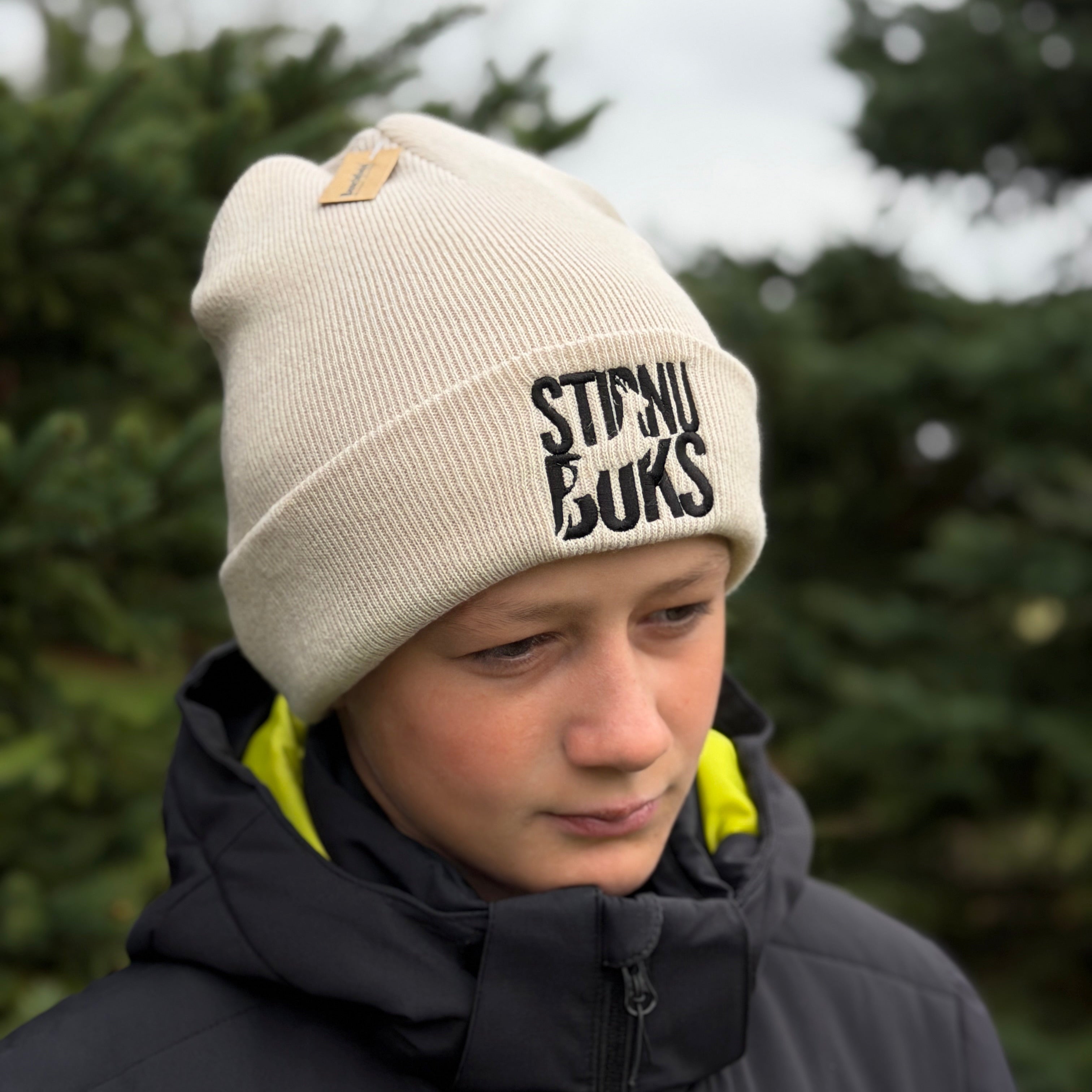 Person wearing a beige beanie with 'STIRNU BUKS' branding outdoors.
