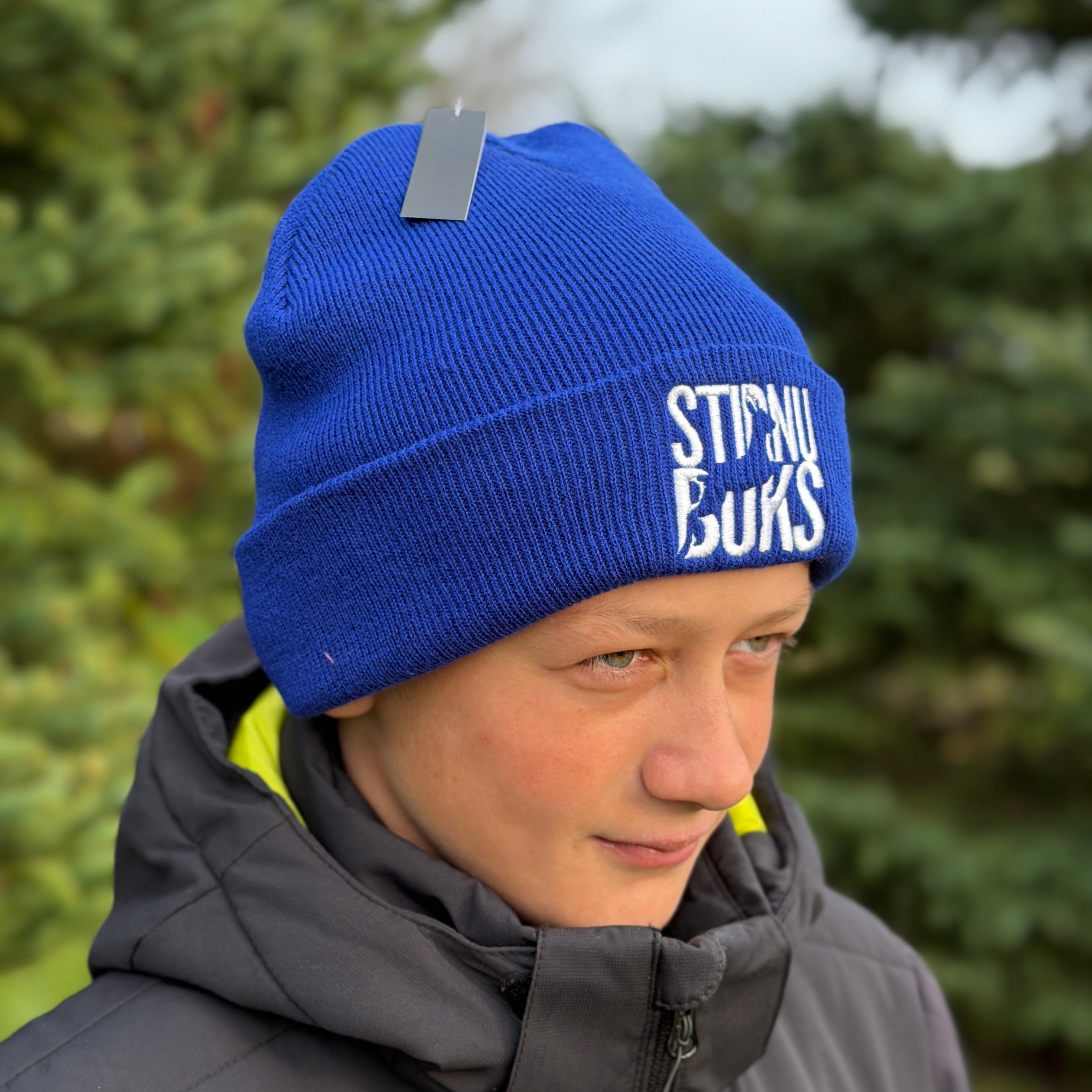 Person wearing a blue beanie with 'Stirnu Buks' logo, standing outdoors with greenery in the background.