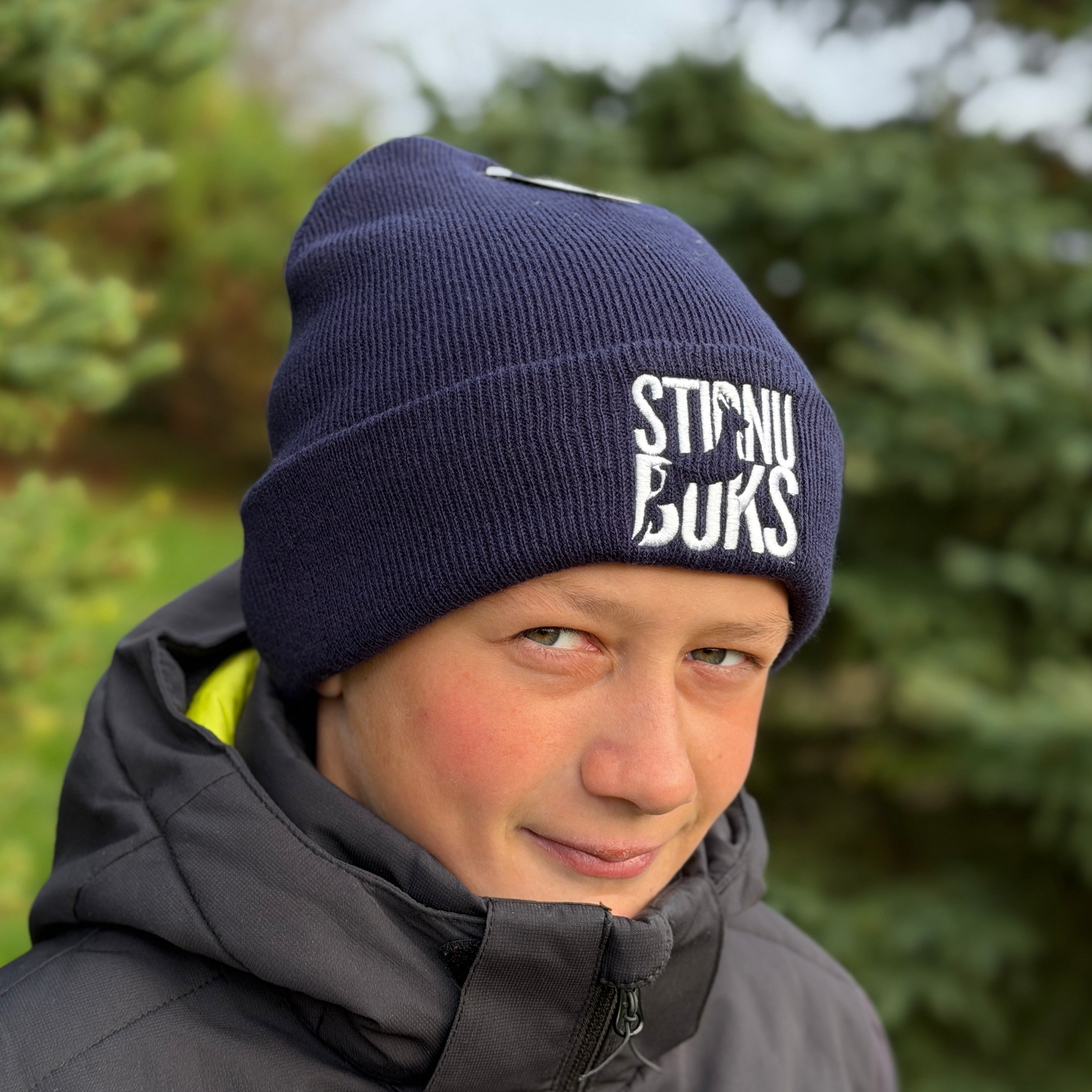 Person wearing a navy blue beanie with 'Stirnu Buks' branding, standing outdoors with greenery in the background.