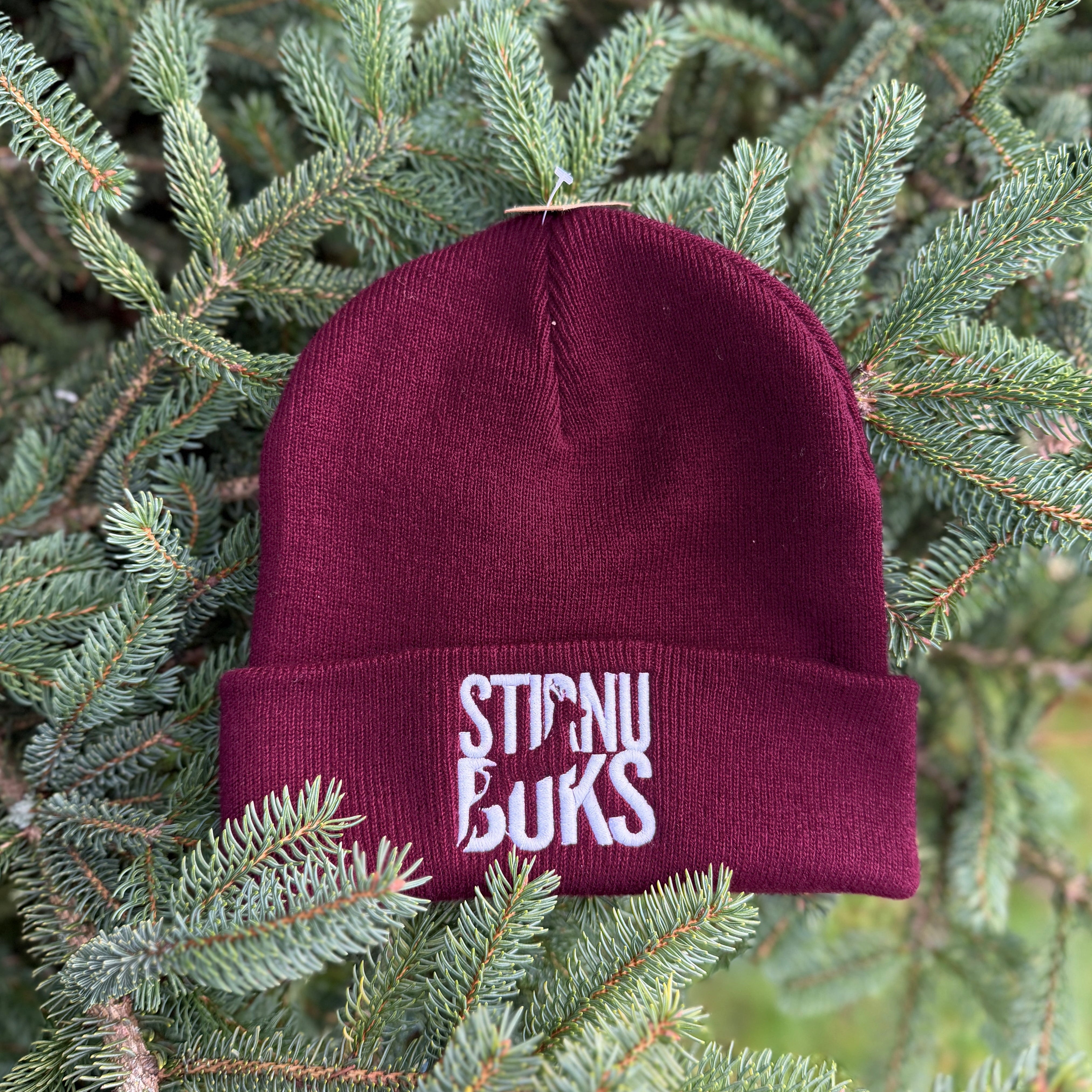 Maroon beanie with 'STIRNU BUKS' text on a green pine branch background