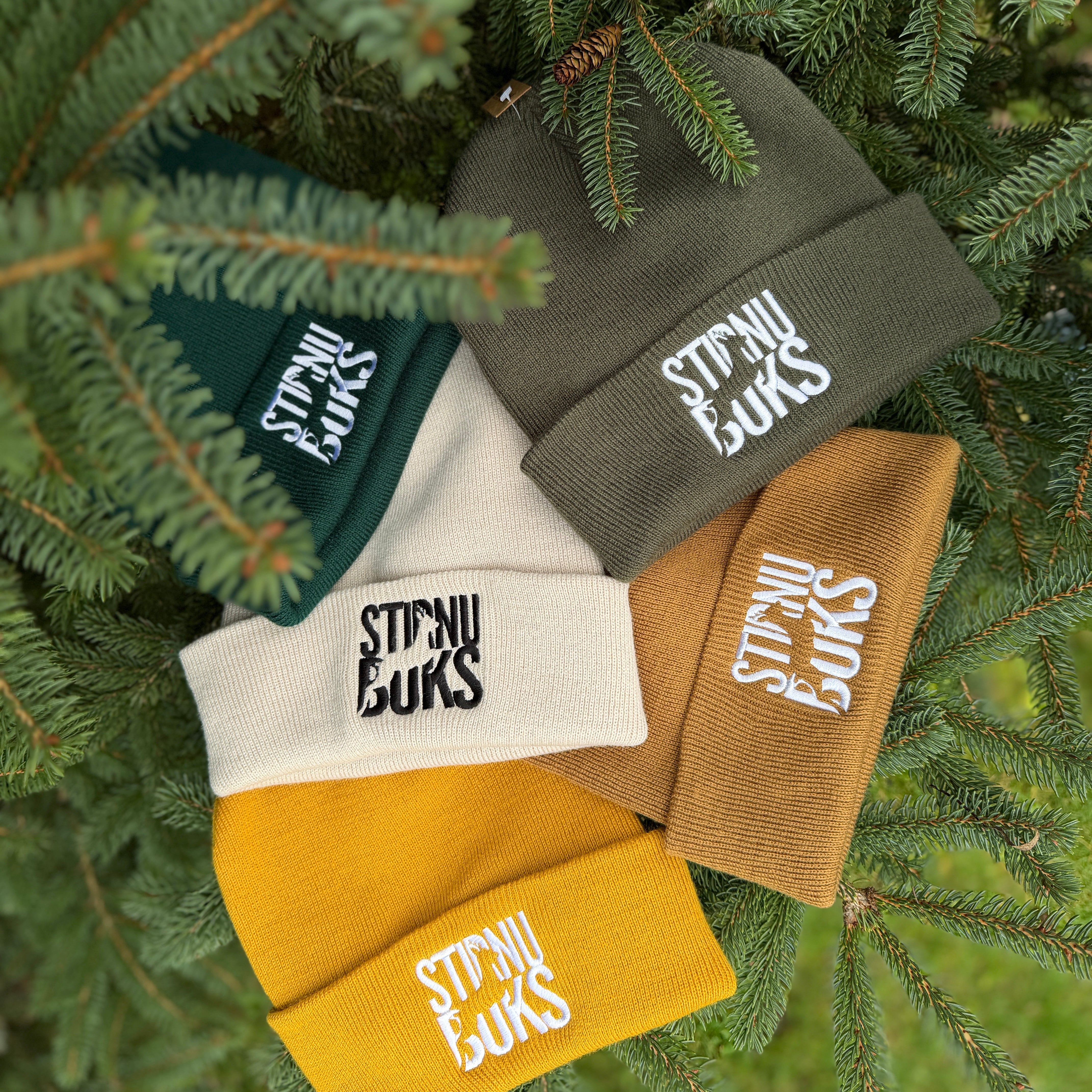 Five beanies in different colors with 'Stirnu buks' branding on a green leafy background