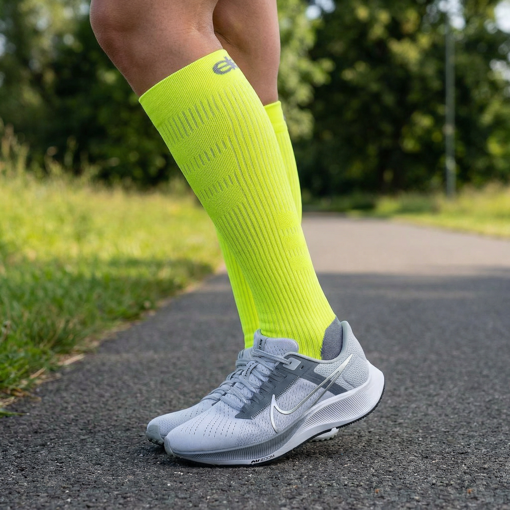Person wearing bright yellow knee-high socks and gray running shoes on a path with greenery.