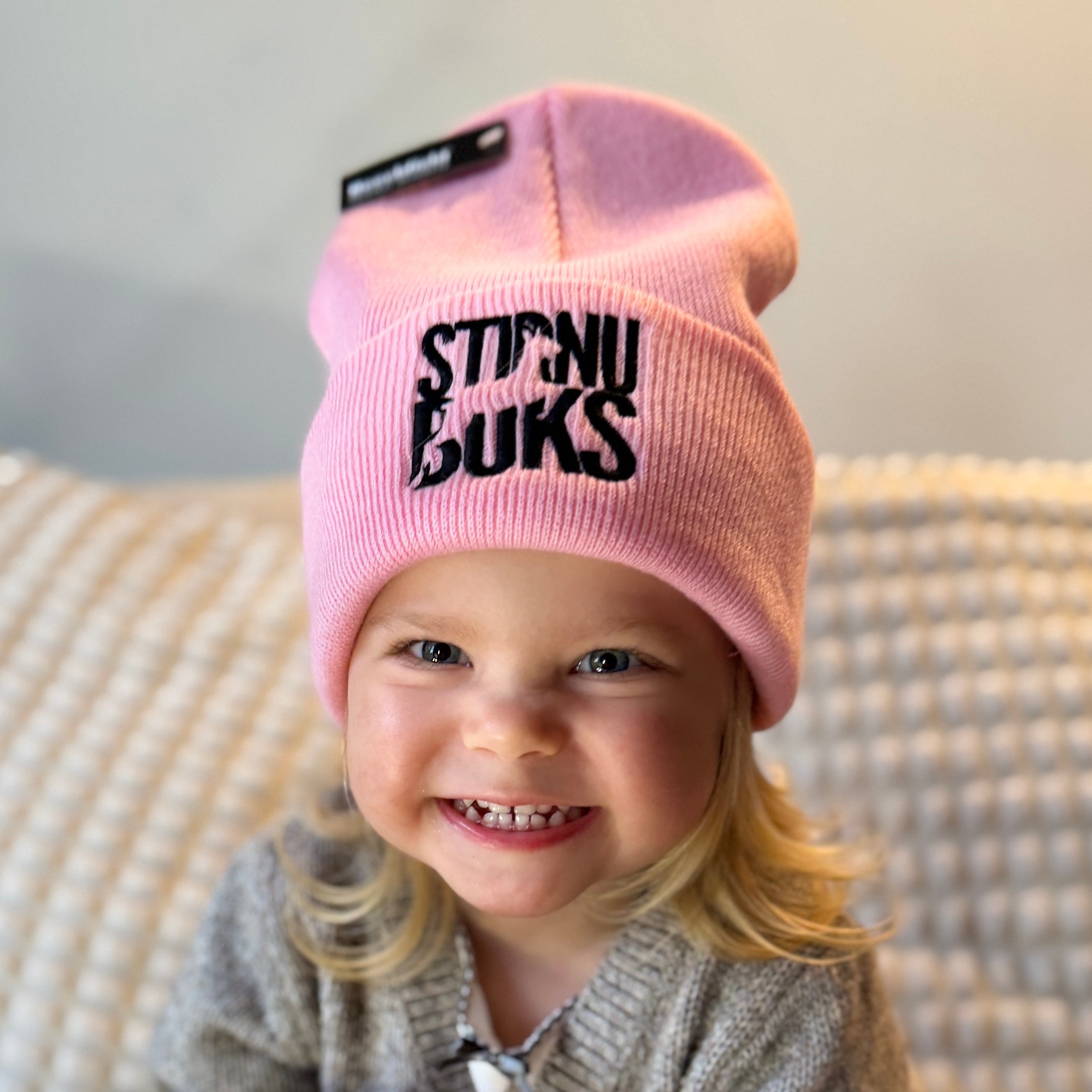 Child wearing a pink beanie with 'STIRNU BUKS' logo, sitting on a textured surface.