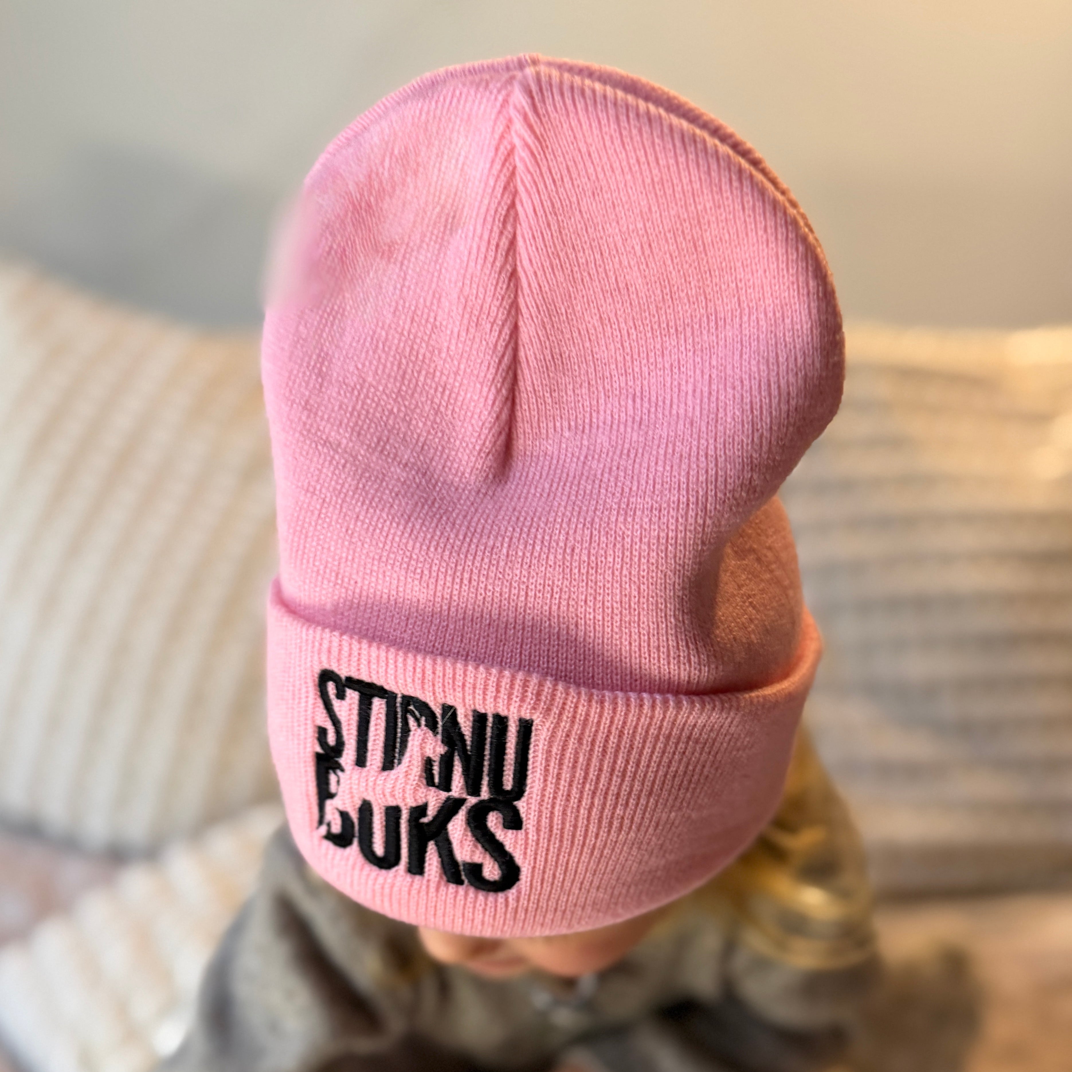 Pink beanie with 'STIRNU BUKS' text worn by a child sitting on a couch.