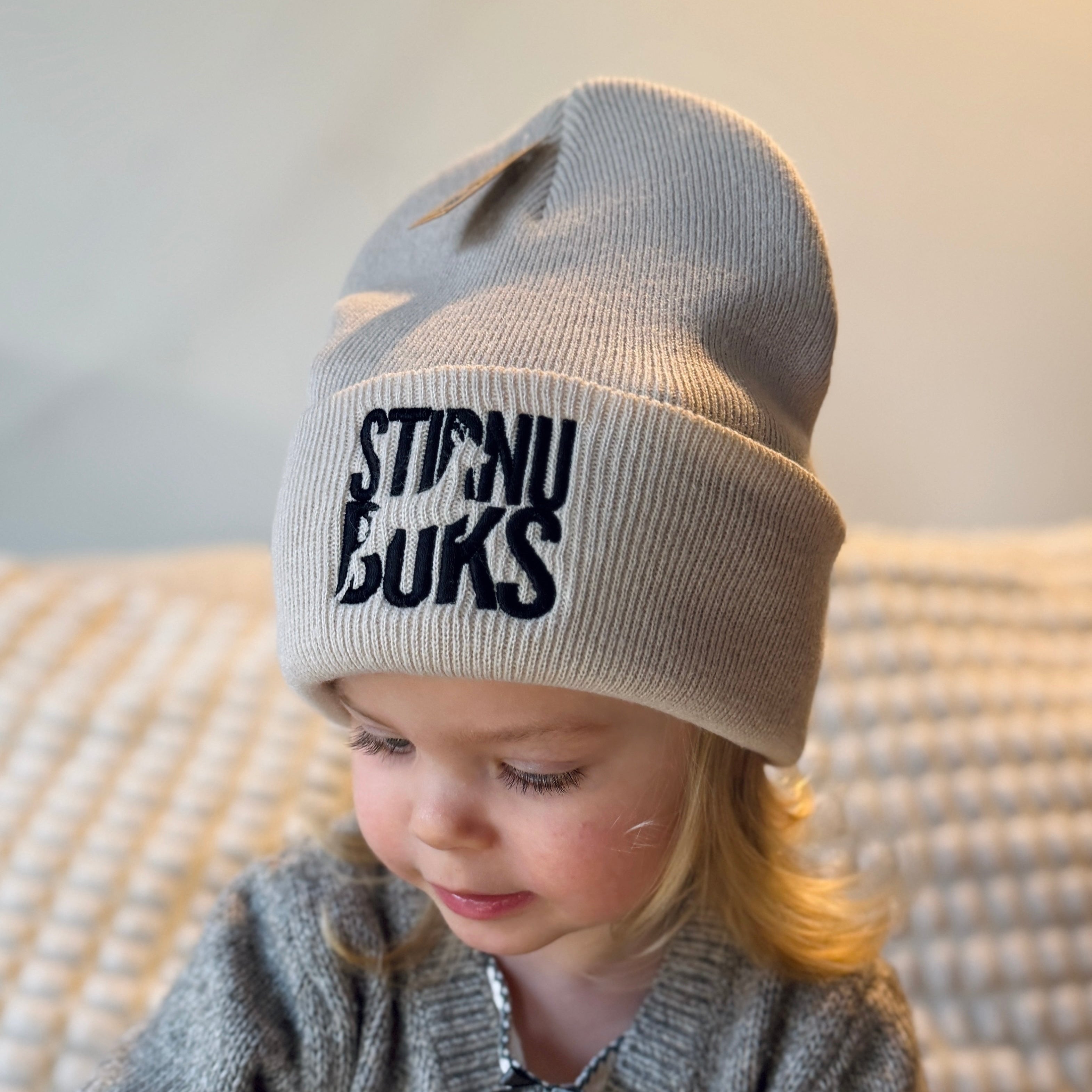 Child wearing a gray knit hat with 'STIRNU BUKS' text, sitting on a textured surface.