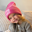 Child wearing a pink beanie with text, sitting on a textured surface.