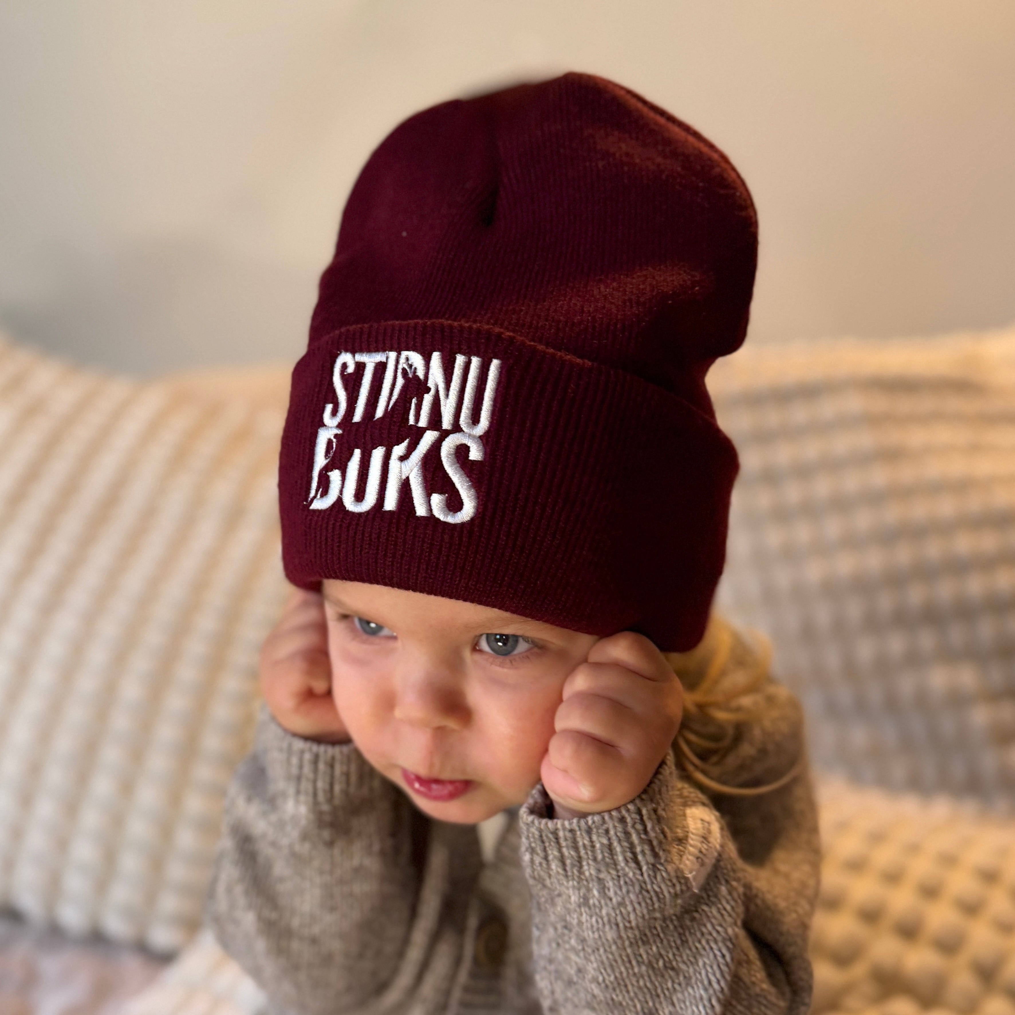 Child wearing a maroon beanie with text, sitting on a textured surface.