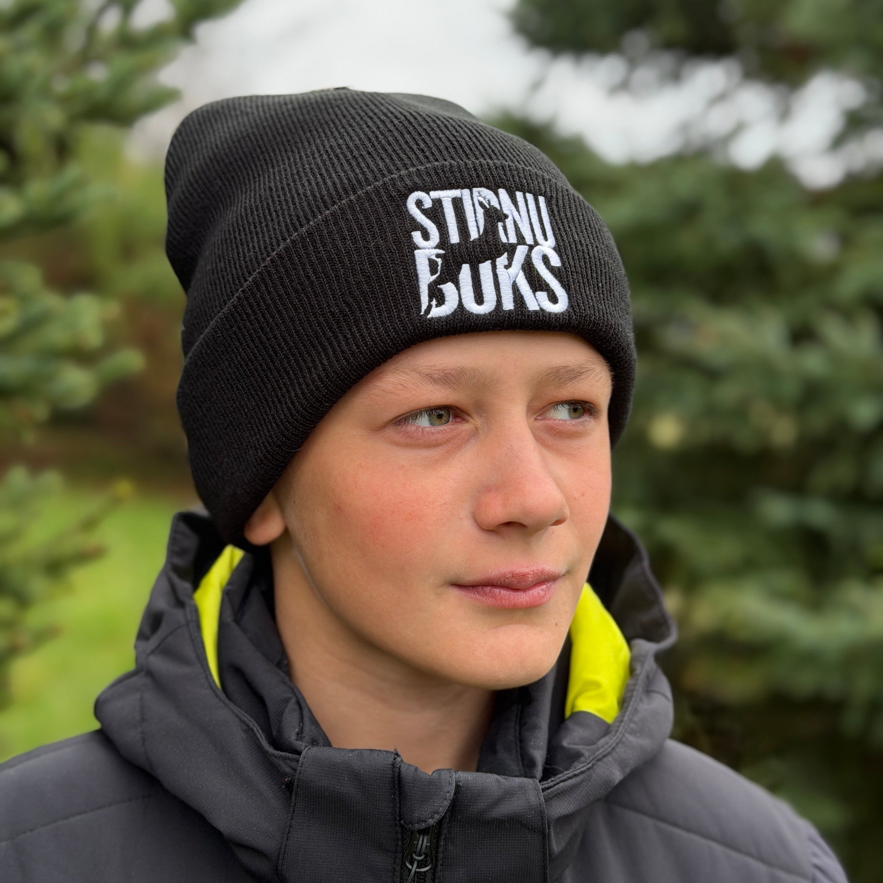 Person wearing a black beanie with STIRNU BUKS logo, standing outdoors with trees in the background.