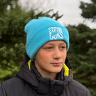 Person wearing a blue beanie with 'STIRNU BUKS' text, standing outdoors with trees in the background.