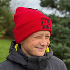 Person wearing a red beanie with 'Stirnu Buks' logo, standing outdoors with trees in the background.