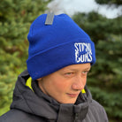 Person wearing a blue beanie with 'Stirnu Buks' logo, standing outdoors with greenery in the background.