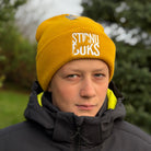 Person wearing a yellow beanie with 'Stirnu Buks' text, standing outdoors with trees in the background.