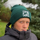 Person wearing a green beanie with 'Stirnu Buks' branding and a dark jacket outdoors.