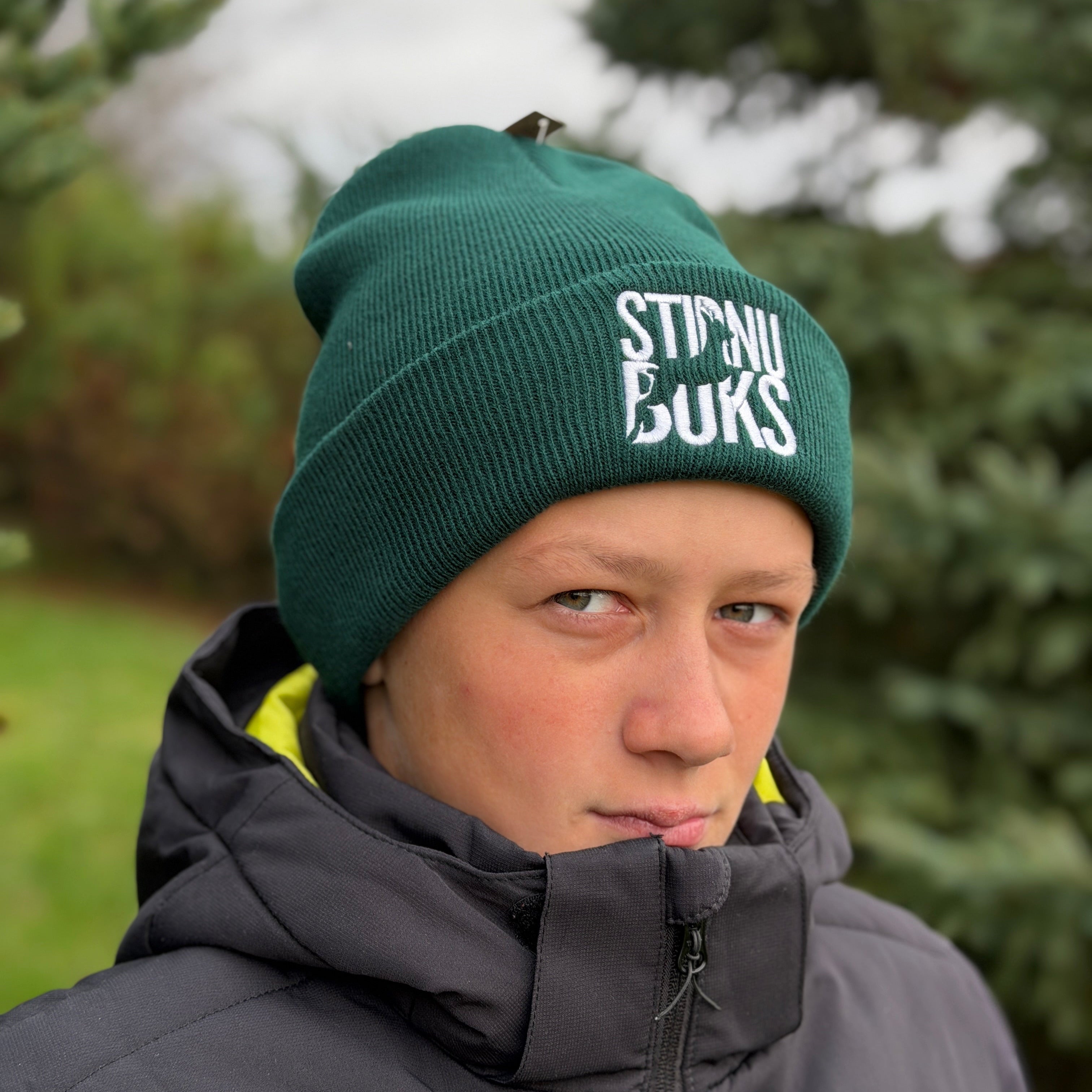 Person wearing a green beanie with 'Stirnu Buks' branding and a dark jacket outdoors.