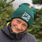 Person wearing a green 'Stirnu Buks' beanie with trees in the background