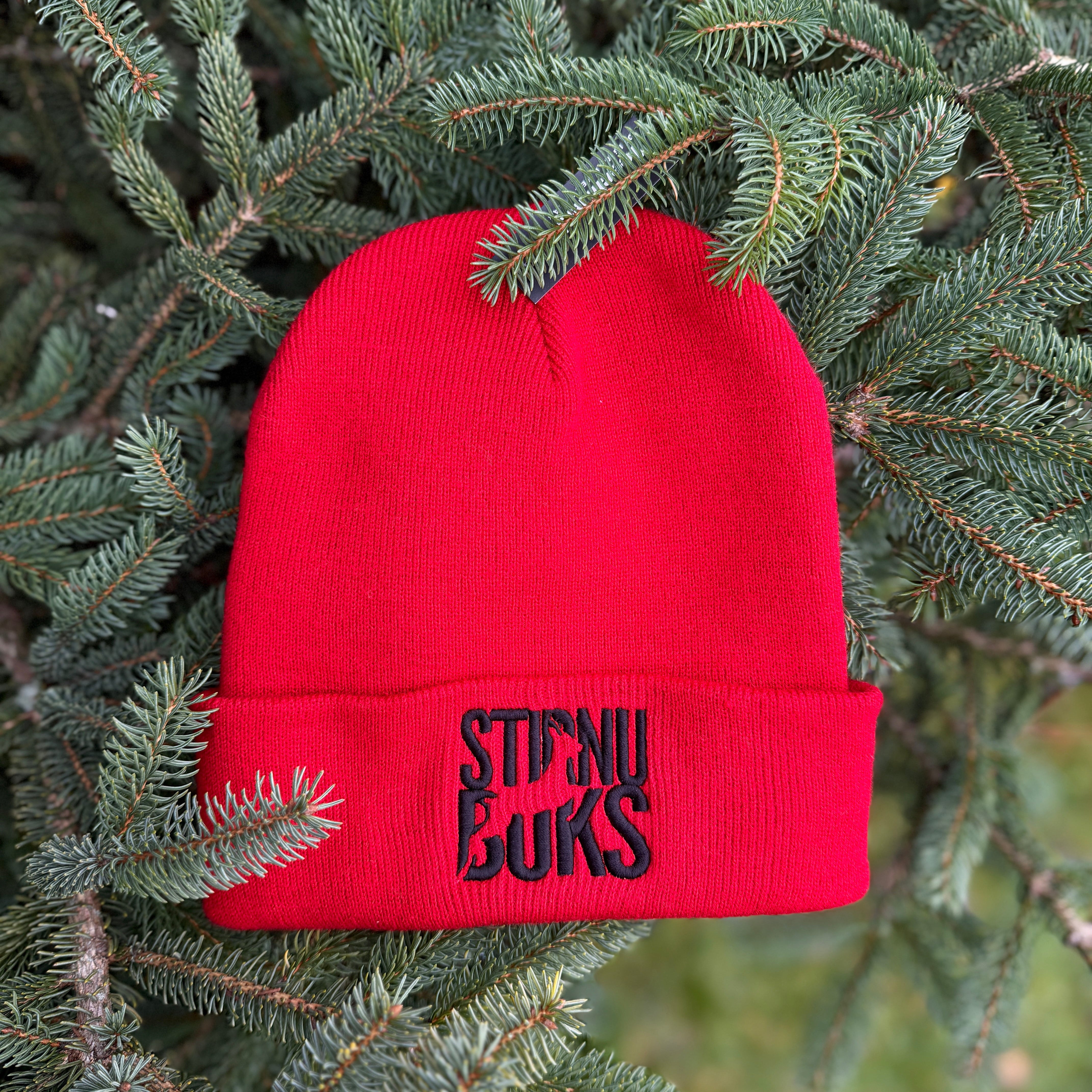 Red beanie with 'STIRNU BUKS' text on a green pine branch background