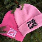 Two pink beanies with 'STIRNU BUKS' branding on a green leafy background