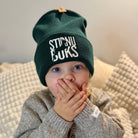 Child wearing a green beanie with text, sitting on a textured surface.