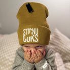 Child wearing a mustard yellow beanie with STIRNU BUKS' logo, sitting on a textured surface.